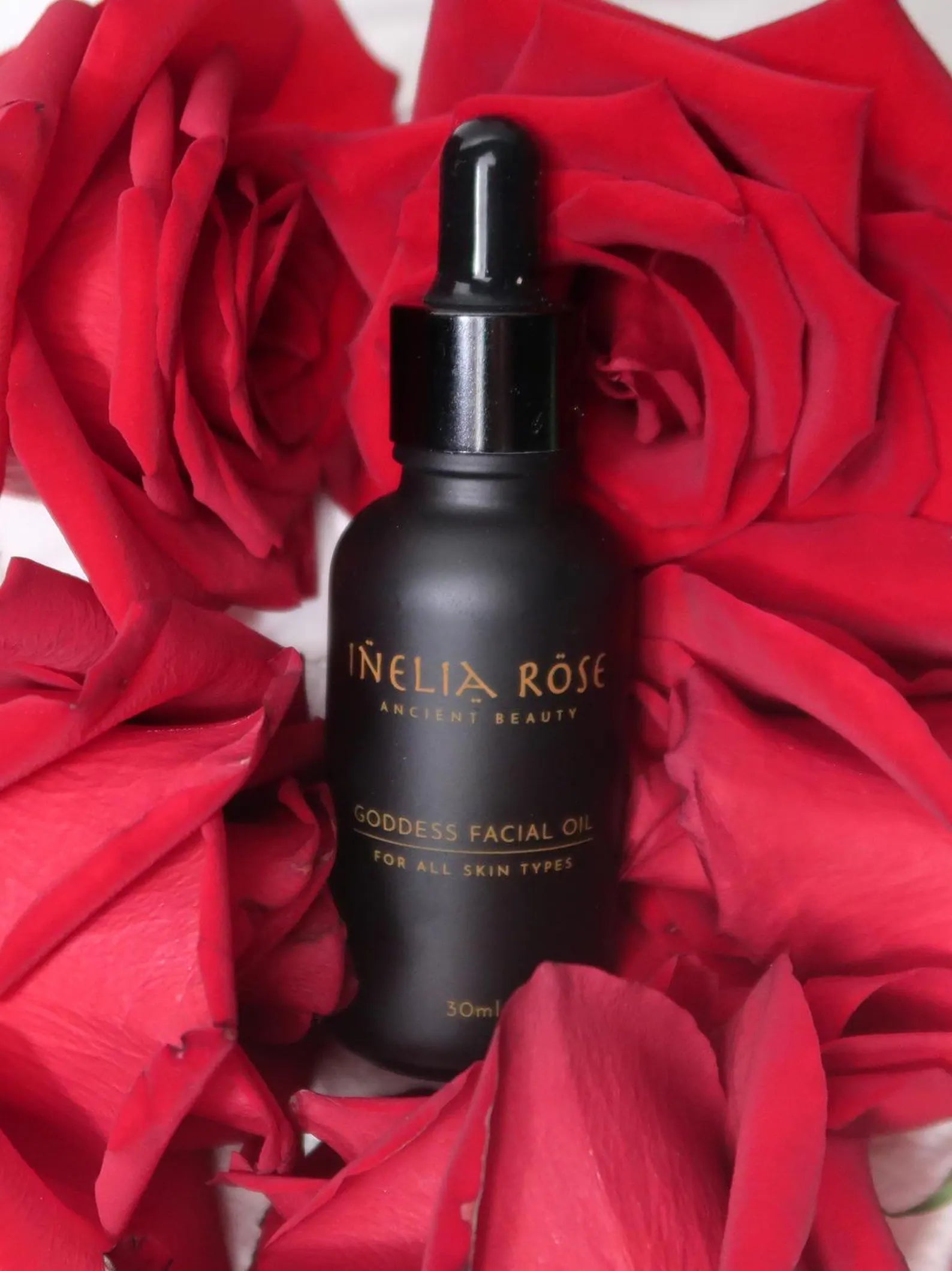Black bottle of Inelia Rose Goddess Facial Oil surrounded by red roses