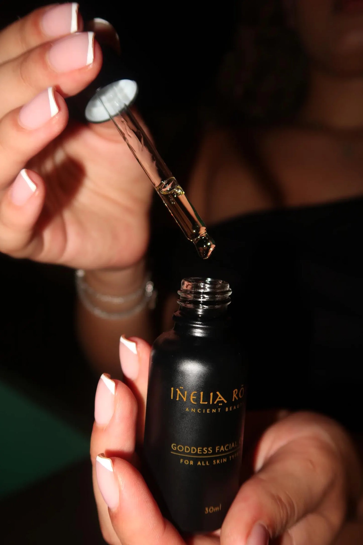 Hand holding a dropper over an Inelia Rara skincare bottle with a dark background